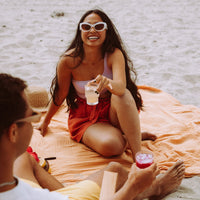 People sitting on a blanket on a beach holding Clevr SuperTeas in glasses with ice.