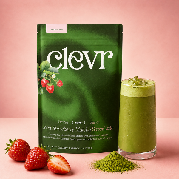 A green stand-up pouch of clevr Iced Strawberry Matcha SuperLatte stands on a pink studio background next to a tall glass of green iced matcha latte, with whole and sliced strawberries and a small pile of green powder in front.